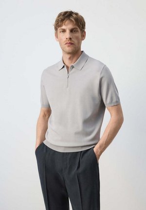 Hombre con cabello castaño claro que lleva una camiseta tipo polo gris clara de manga corta con cremallera y pantalones oscuros, de pie con las manos en los bolsillos.