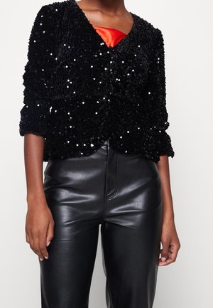 Sort palietter cropped jakke over rød top kombineret med høj taljede sorte læderbukser på en stående person.