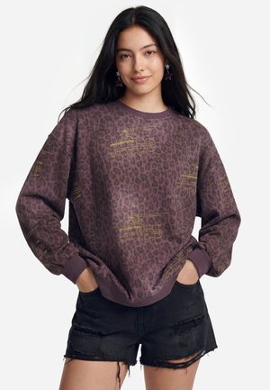Ung kvinde iført en lilla leopardprint-sweatshirt med gul tekst og sorte slidte denimshorts, hænderne i lommerne, smilende let.