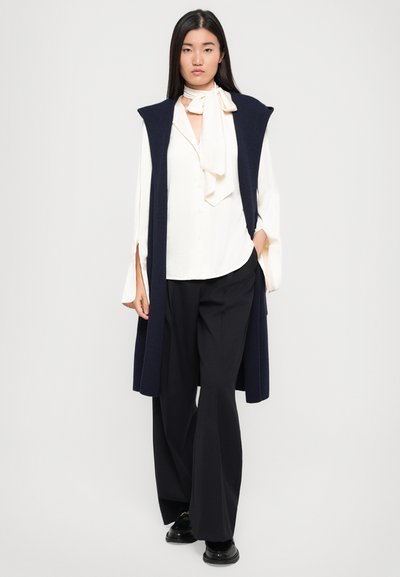 Mornarsko pleteno brezrokavnik z odprtimi stranicami preko bele bluze z zavezanim ovratnikom. Črne široke hlače s črtami. Črni čevlji z sijočo površino.