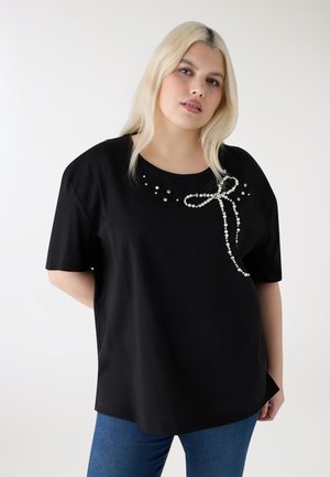 Donna bionda che indossa una maglietta nera a maniche corte decorata con un fiocco di perle vicino allo scollo e jeans blu, in piedi davanti a uno sfondo bianco.