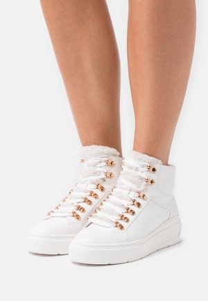 Bílé vysoké tenisky se zlatými očky a bílými tkaničkami, nošené na bosých nohou, s plyšovou podšívkou u kotníku.