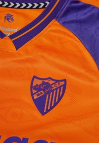 Orange sportströja med lila axel detaljer, rundad hals, präglade mönster och en blå sköldlogotyp med "MALAGAC.F."