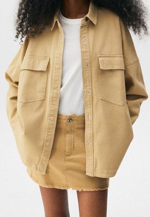 Femme portant une veste surdimensionnée couleur beige à boutons et une mini-jupe en denim effiloché assortie, par-dessus un haut blanc, les mains dans les poches de la veste.