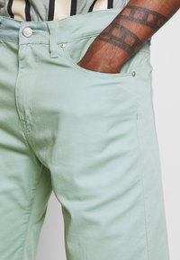 Dark-skinned arm with filmstrip tattoo resting in front pocket of mint green pants with visible button and stitching details.
