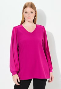 Blusa fucsia con scollo a V, maniche lunghe a pieghe e polsini aderenti. Tessuto morbido con una trama liscia, abbinata a pantaloni neri.