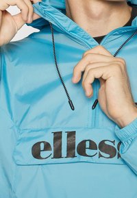 Světle modrý větrovka s designem na půl zip, nastavitelnou kapucí se stahovacími šňůrkami a velkým černým logem "ellesse" na přední straně.