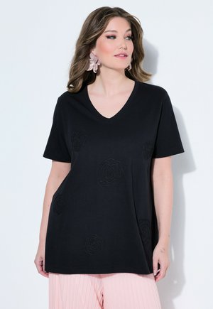 Vrouw die een zwart V-hals shirt met korte mouwen draagt met subtiele rozenpatronen en lichtroze geplooide broek, licht naar rechts gericht.