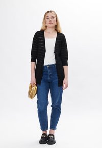Cardigan a righe nere sopra una canotta bianca, abbinato a jeans blu. Borsa dorata e mocassini neri con dettaglio a catena. Trame morbide.