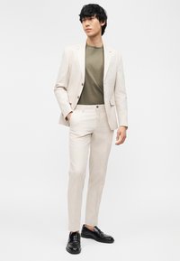 Νέος άντρας που φοράει ανοιχτό μπεζ κοστούμι με ταιριαστό παντελόνι, ελιά πράσινο μπλουζάκι και μαύρα δερμάτινα loafers, στέκεται με το ένα χέρι στην τσέπη.