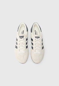 Λευκά σουέντ αθλητικά παπούτσια με μαύρες δερμάτινες ρίγες, στρογγυλή μύτη και επίπεδη λαστιχένια σόλα. Διαθέτουν λευκά κορδόνια και επένδυση με το εμπορικό λογότυπο στο εσωτερικό.