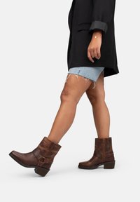 Stivali alla caviglia in pelle marrone con punta tonda, tacco a blocco e dettagli con cinturini. Indossati con pantaloncini di jeans chiari e un blazer nero oversize.