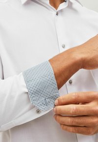 Biely košeľový dres s textúrovaným povrchom, modré vzorované detaily na manžetách a gombíkovými manžetami. Ruka upravuje manžetu, pričom zdôrazňuje dizajnové prvky.
