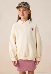 Sweatshirt crème avec un col, patch en forme de cœur et manches bouffantes, associé à une jupe en velours côtelé rose. Casquette en imprimé léopard brun.