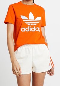 Orange bomullströja med en vit Adidas-logotyp och tre-stripesdesign. Matchas med vita shorts som har en liten röd Adidas-logotyp.