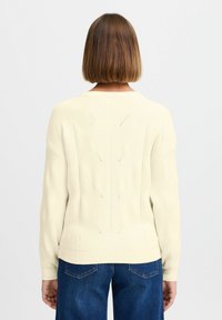 Krémový svetr s ribkovanou texturou, s kulatým výstřihem a geometrickými vzory na zadní straně. Nesený s modrými džíny.