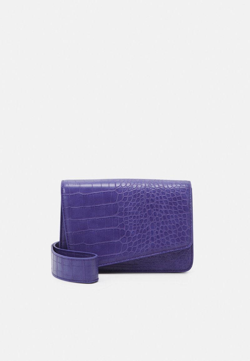 Sac bandoulière rectangulaire violet avec texture crocodile et large sangle enroulée à côté sur un fond clair.