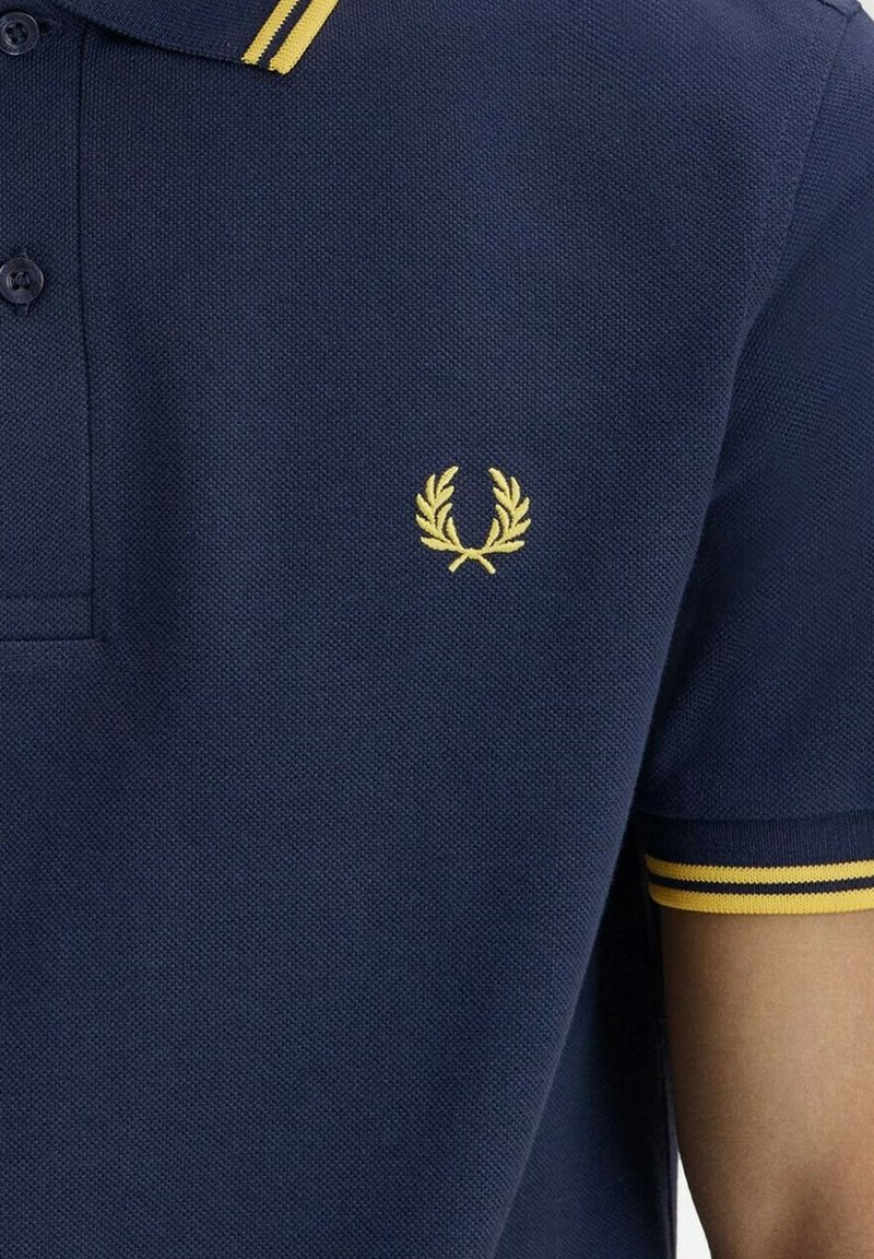 Polo azul marino con logo de corona de laurel amarilla en el pecho y doble franjas amarillas en el cuello y el puño de la manga.