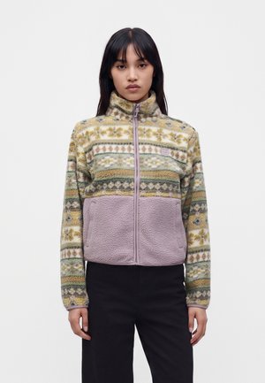 Young woman wearing a high-neck zip-up fleece jacket with floral and striped patterns in green, yellow, and lavender tones, and black pants.