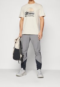 Beige tričko s krátkým rukávem s grafikou hory a slunce, šedé lehké kalhoty a dvoubarevný batoh v krémové a černé barvě. Šedé tenisky.