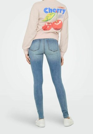 Nainen, jolla on päällään vaaleansiniset kapeat farkut, valkoiset tennarit ja beige collegepaita, jossa on suuret kirsikkakuviot ja takana teksti "Cherry".