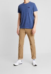 Granatowa koszulka z krótkim rękawem o teksturowanej tkaninie i małym logo na lewej piersi, zestawiona z beżowymi chinosami i białymi trampkami.