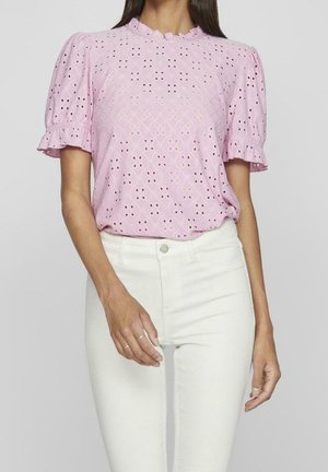 Femme portant un haut en dentelle violet clair à manches courtes avec des détails œillets et un pantalon blanc taille haute, debout sur un fond uni.