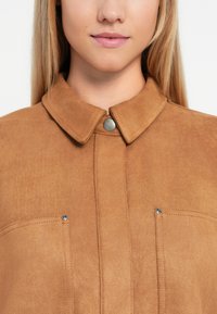 Brown suede jacket featuring a collar, snap button closure, and two chest pockets with small metal accents.