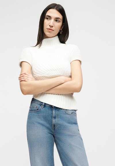 Γυναίκα με ίσια σκούρα μαλλιά που φοράει λευκό ριμπ πουλόβερ με κοντά μανίκια και μπλε τζιν, με σταυρωμένα τα χέρια, κοιτώντας προς τα πλάγια.