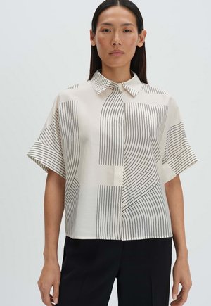 Frau mit langen dunklen Haaren trägt ein weißes Kurzarmshirt mit schwarzen geometrischen Streifen und schwarze Hosen und steht vor einem schlichten hellen Hintergrund.