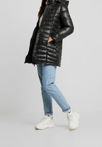Černá, nadýchaná péřová bunda s kapucí a zipem, s prošívaným designem. V kombinaci se světle modrými džíny a bílými teniskami.