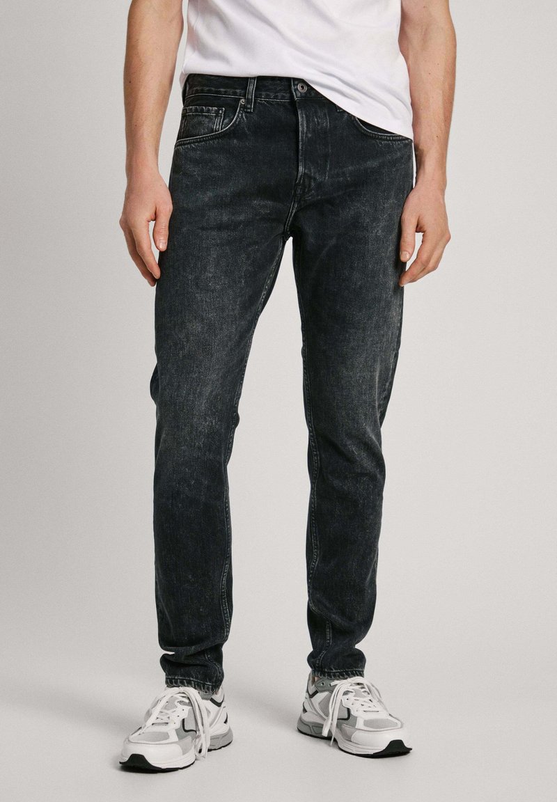 Slim-fit jeans w kolorze ciemnego denimu, wykonane z denimu, z klasycznym pięciopocketowym fasonem i subtelnymi przetarciami. Noszone z białymi butami sportowymi.