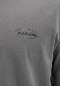 Camiseta de algodón gris con un logo redondo negro en el pecho que contiene el texto "Jack & Jones Club 1990" dentro de un contorno ovalado.