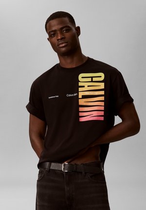 Άνδρας που φοράει μαύρο T-shirt Calvin Klein με μεγάλο κατακόρυφο κείμενο με βαθμιαία μετάβαση χρωμάτων και μαύρα τζιν, στέκεται μπροστά σε απλό γκρι φόντο.