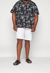 Svart t-shirt med kort ärm och vitt blommönster, kombinerat med vita uppvikta shorts och svarta slidesandaler. Slät tygtextur.