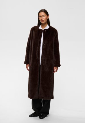 Femme debout portant un long manteau en fausse fourrure marron foncé sur une chemise blanche, un pantalon noir et des chaussures noires sur un fond uni.