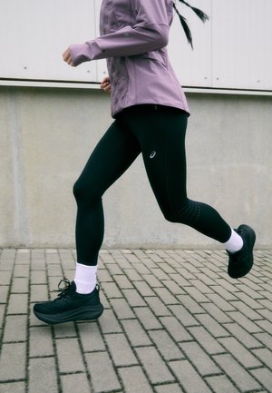 Leggings atléticos pretos e uma jaqueta lilás clara. As leggings têm um padrão subtil perto das panturrilhas, combinadas com sapatilhas pretas e meias brancas.