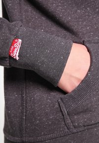 Sudadera con capucha de color gris oscuro con una tela texturizada. Presenta un bolsillo frontal y puños acanalados. Incluye un parche con el logo rojo "SuperDry" en la manga.