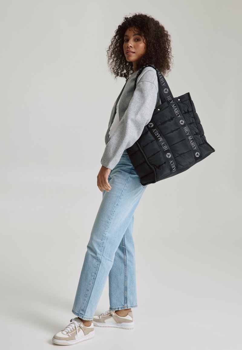 Zwarte, gewatteerde tas met logo-geprinte bandjes, met een rechthoekige vorm en gewatteerde textuur. Draagtas over de schouder met denim jeans.