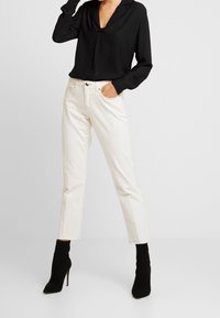 Blouse noire à manches longues avec un col en V, associée à un jean droit écru avec des ourlets effilochés et des bottines à talons hauts noires.