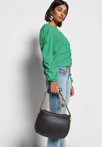 Schwarze Handtasche aus Kunstleder mit geschwungener Form und Kettenriemen, verziert mit einem goldenen Logo und einer glatten Oberfläche, gehalten von einer Person in Jeans und einem grünen, gestreiften Oberteil.