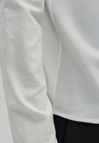 Haut blanc à manches longues avec un tissu texturé d'un côté, une finition lisse de l'autre, et un panneau noir contrastant en bas.