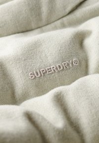 Lys beige stof med en blød tekstur prydes af det broderede "SUPERDRY" logo i lys tråd, hvilket viser et subtilt herringbone mønster.