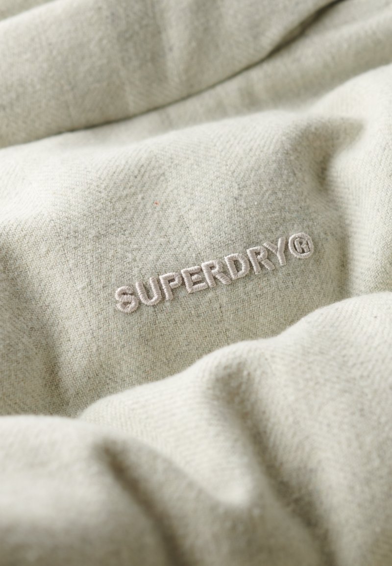 Lichtbeige stof met een zachte textuur kenmerkt zich door het geborduurde "SUPERDRY"-logo in een lichte draad, met een subtiel visgraatpatroon.
