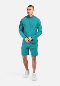 Türkise Zip-Jacke und passende Shorts aus glattem Stoff. Weiße Sneakers runden das Outfit ab. Schlichtes, modernes Sportdesign.