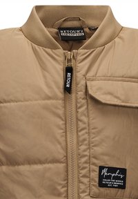Beige Puffer-Jacke mit geripptem Kragen, Reißverschluss vorne und Tasche auf der linken Brust. Sie bietet Abzeichen und eine glatte, glänzende Textur.