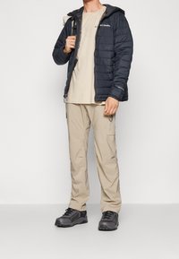 Męski strój na zewnątrz: granatowa puchowa kurtka, beżowa koszulka, beżowe spodnie typu cargo i szare buty trekkingowe. Elementy wyposażenia to kaptur i kieszenie na suwak.