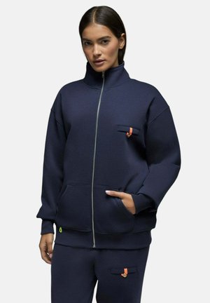Femme portant une veste zippée bleu marine et un pantalon assorti avec de petits boutons orange, debout avec une main dans la poche.
