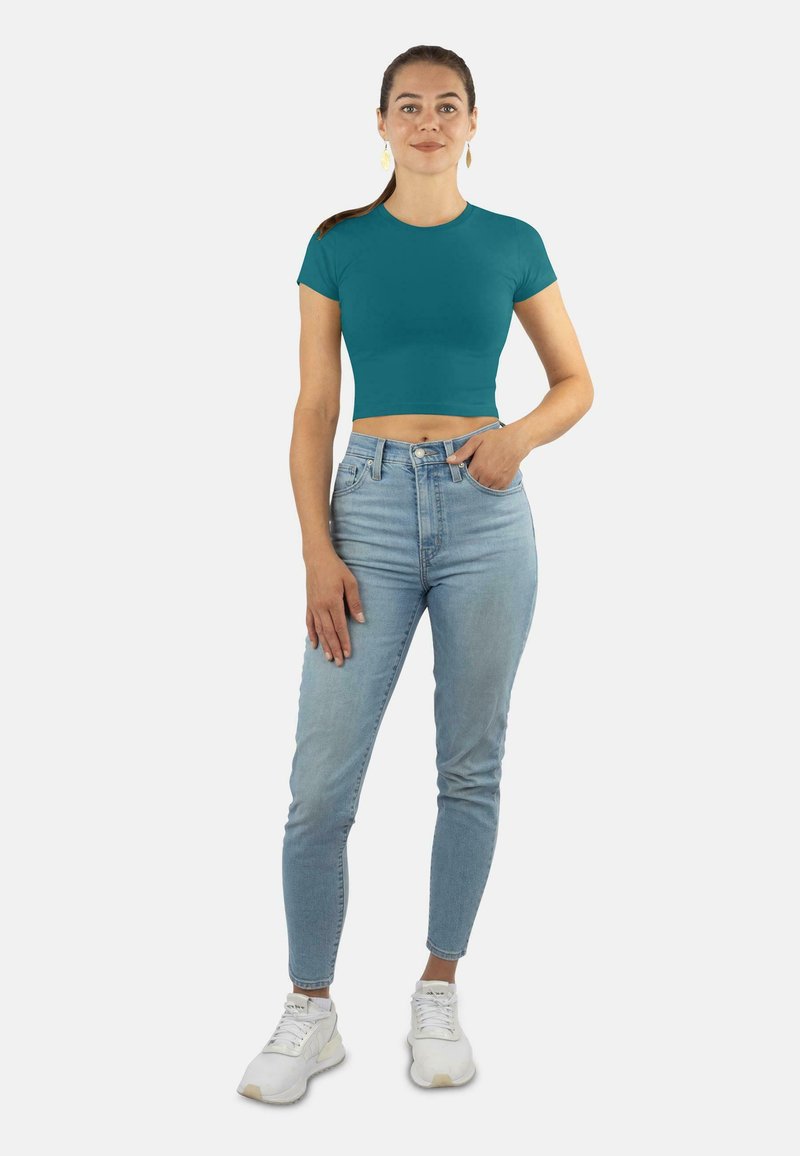 Türkisfarbenes, kurzes T-Shirt mit kurzen Ärmeln, kombiniert mit hellblauen High-Waist-Jeans und weißen Sneakers. Die Hauttextur ist sichtbar.
