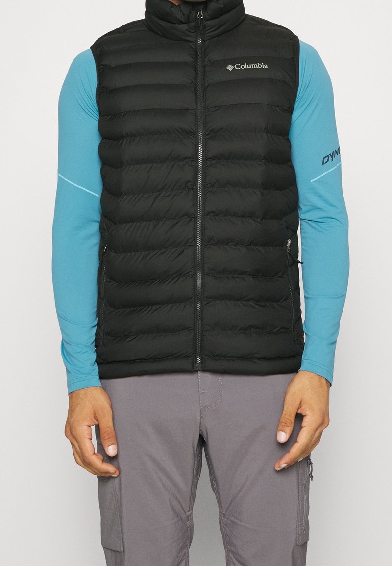 Gilet noir isolé avec un col montant, fermeture éclair à l'avant et poches latérales. Porté par-dessus un haut à manches longues bleu, avec un tissu texturé.